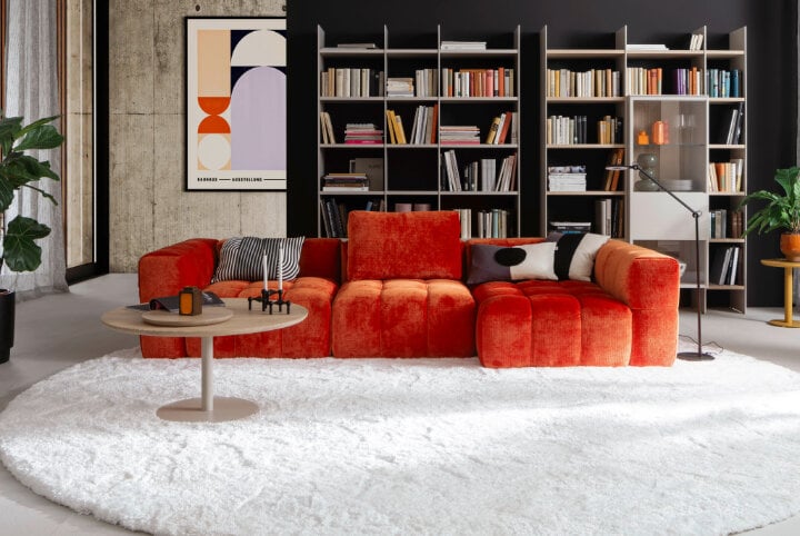 Wohnzimmer mit Dreisitzer Sofa, links ein großes Sessel, rechts ein großes helles fenster und auf dem Boden ein dicke Teppich ausgelegt